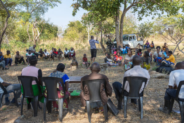 Community members, traditional leaders, and conservation representatives gather for an outdoor meeting under trees in Zambia, sitting in a large circle.