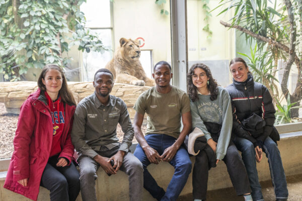 Five Frankfurt Spring School participants, Jana Scherbarth, Sturburn Chuma, Gift Chambatu, Johanna Mitra, and Céline Partridge, sit together in front of a lion enclosure at Frankfurt Zoo.