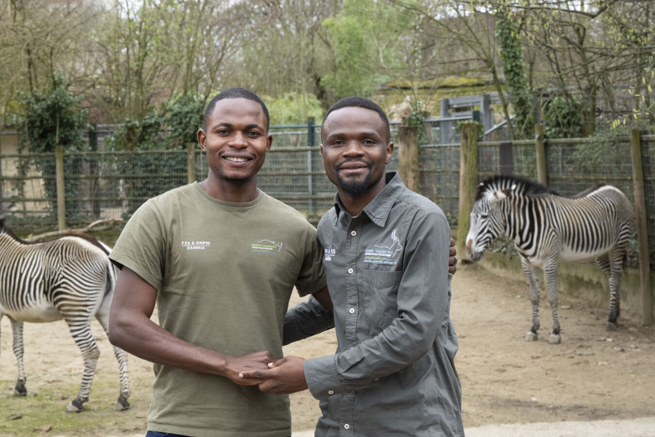 Gift Chambatu (left) and Sturburn Chuma (right), two conservationists from Zambia, standing together at Frankfurt Zoo with zebras in the background.
